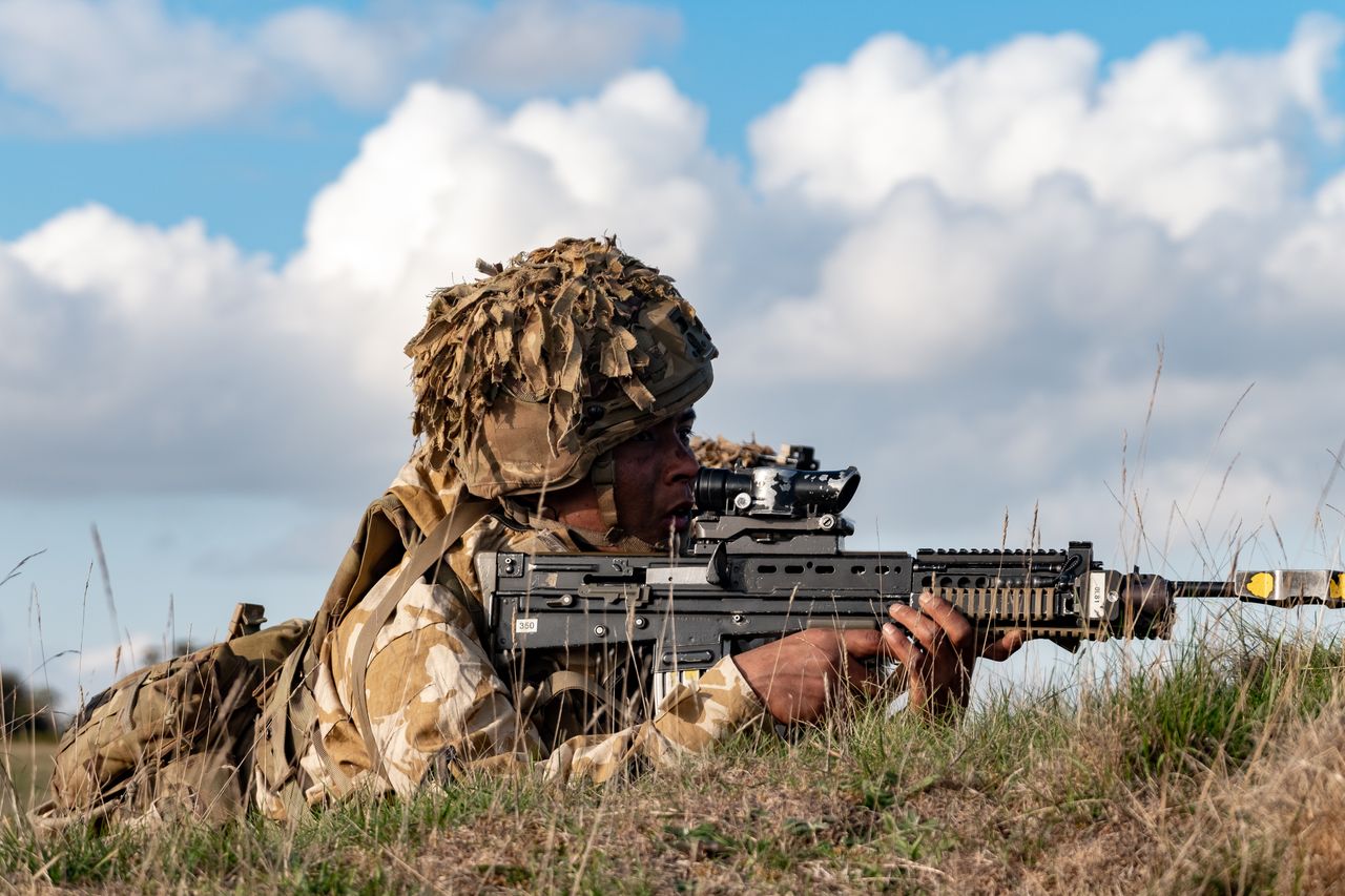 A soldier in camouflage uniform aims a rifle while lying in a grassy field under a blue sky.
